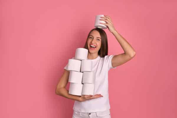 Laughing adult brunette girl in good spirits holding a lot of toilet paper rolls, has a happy look, putting one toilet paper roll onto her head, has fun, she is prepared for a lockdown due Covid-19